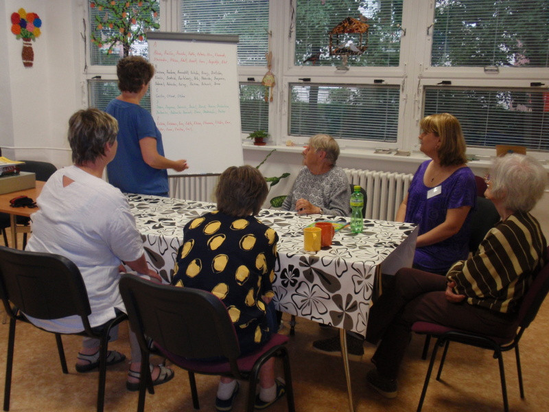denní stacionář s pacientkami sedícímí kolem stolu v úhledně upravené místnosti, jedna osoba předává u flipchartu informace ostatním